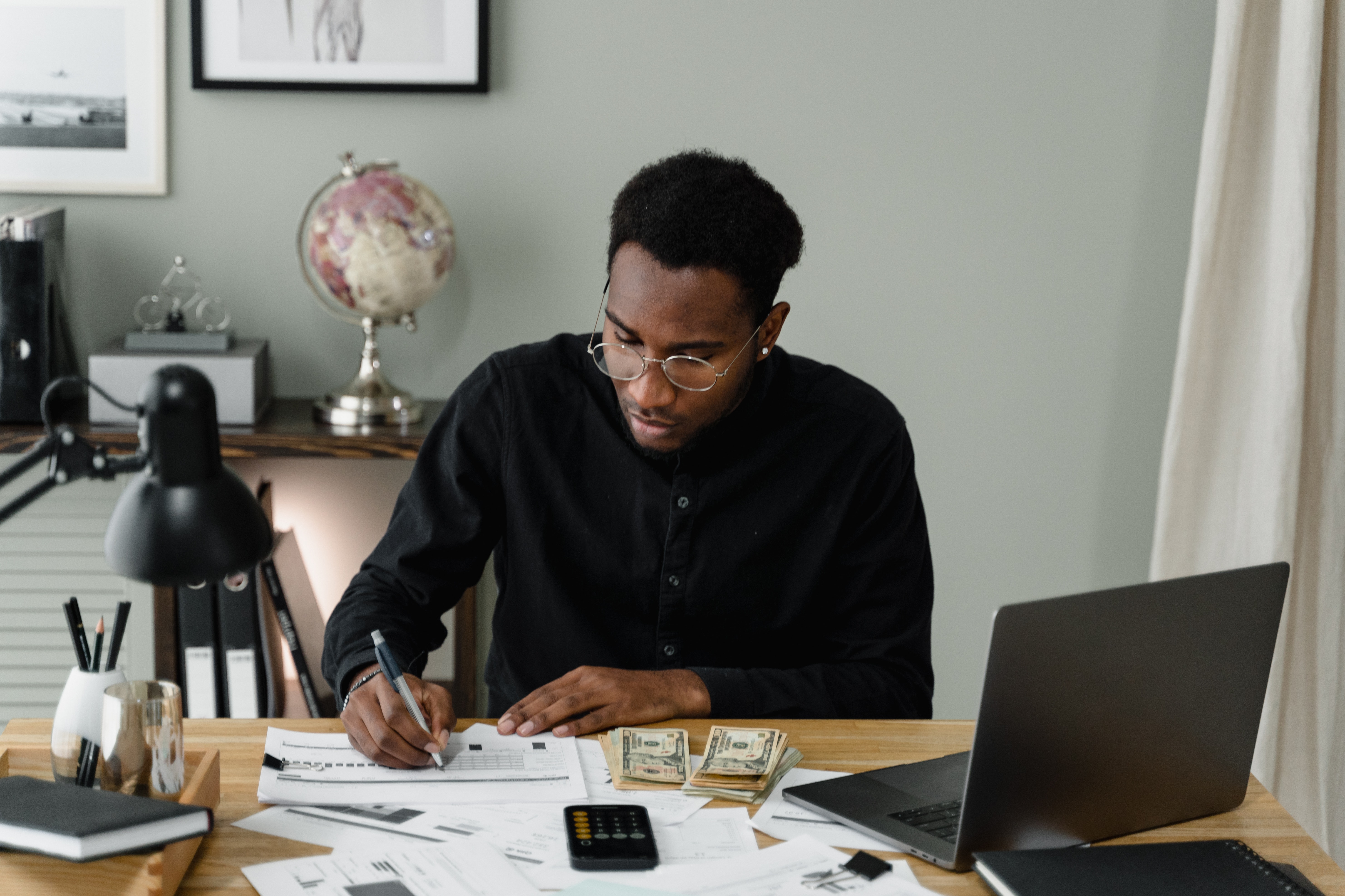 A man writing at a desk with cash and a laptop.