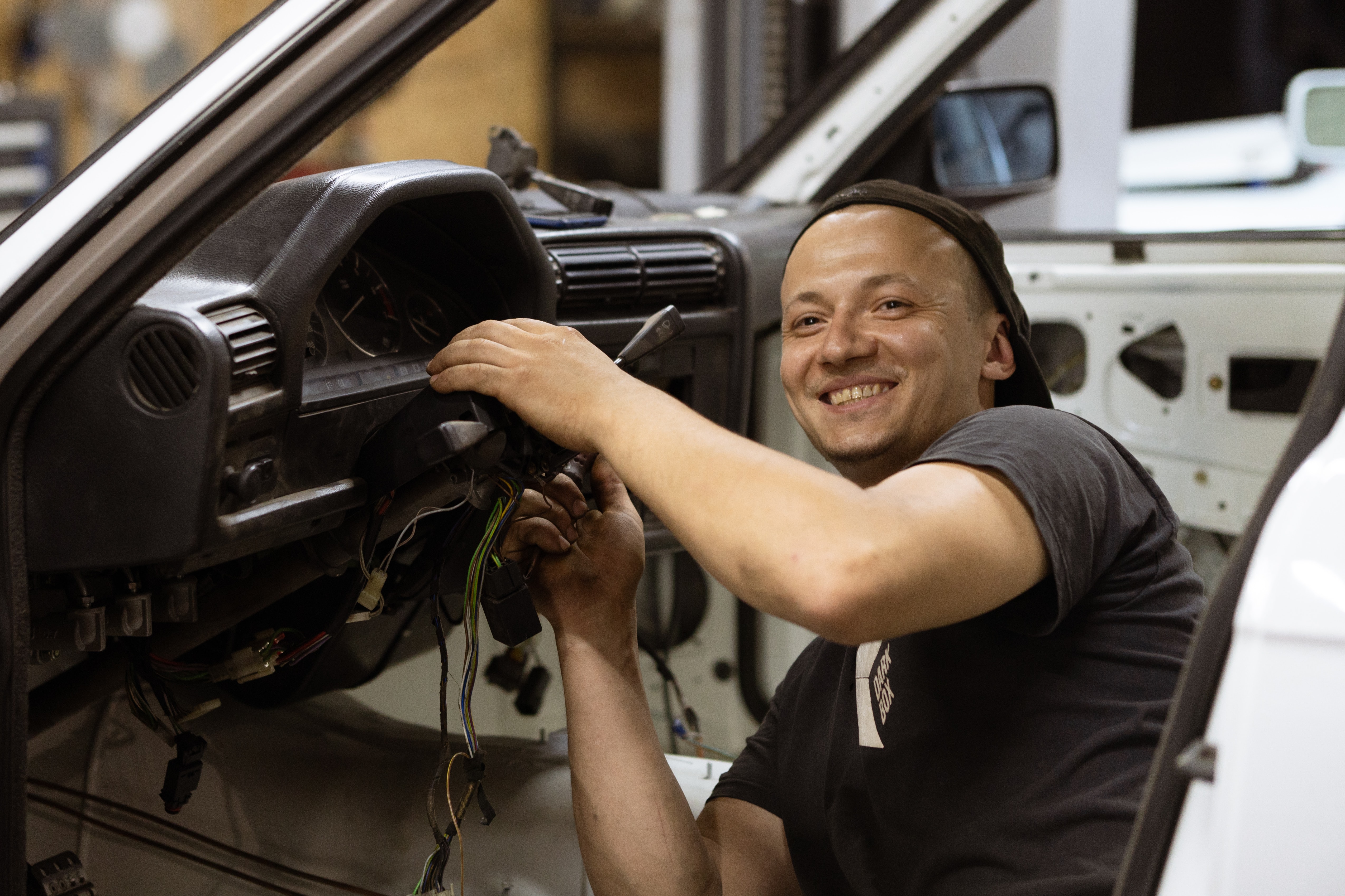 A photo of a automotive mechanic working underneath the dashboard of a car who has stopped for a moment to smile at the camera a have his picture taken.