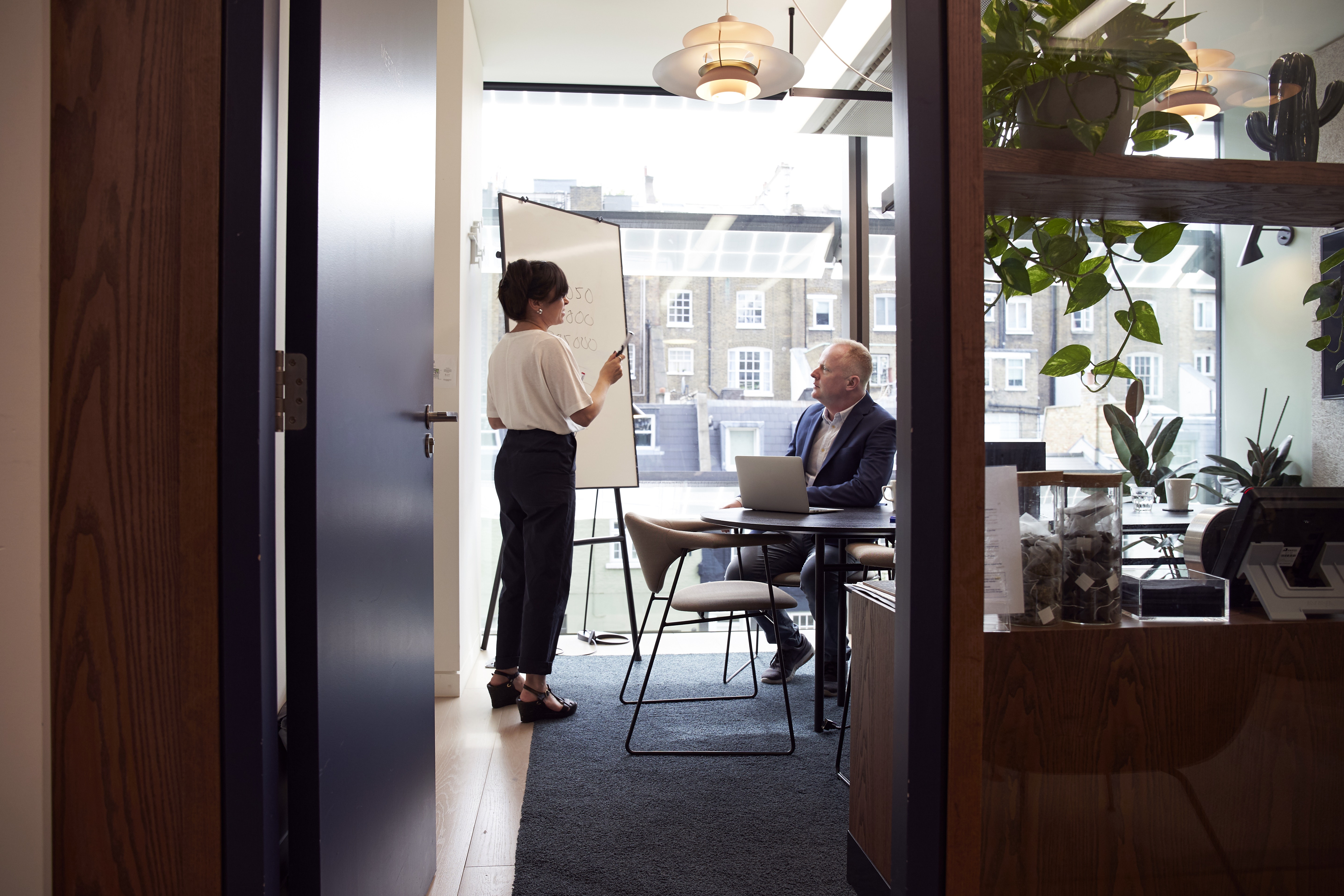 A woman standing at a whiteboard, speaking to a man seated in front of her.