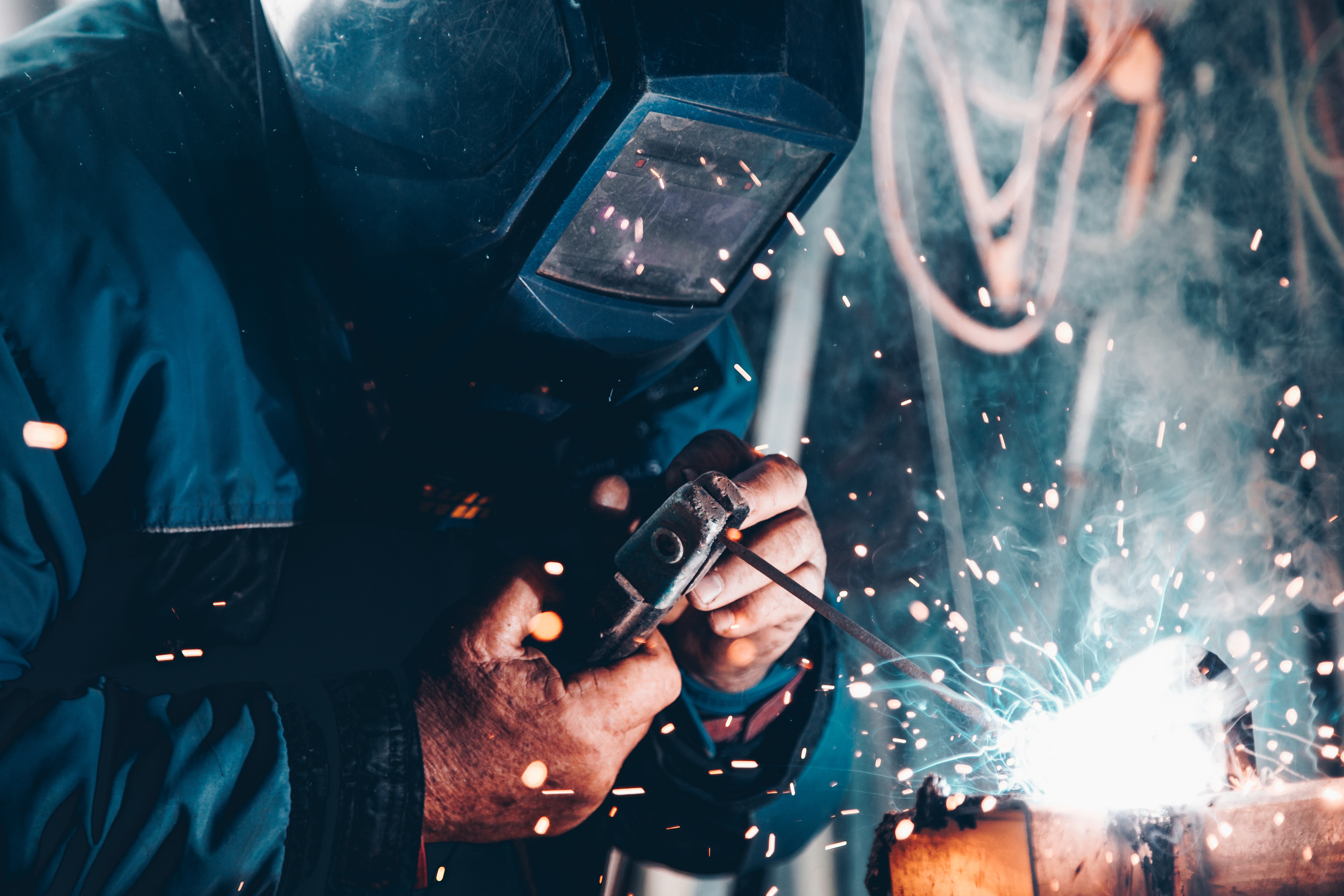 A closeup photo of a man welding with his welder's face mask on, who looks like he's in blue coveralls. The bright light of his arc welding can be seen closeup.