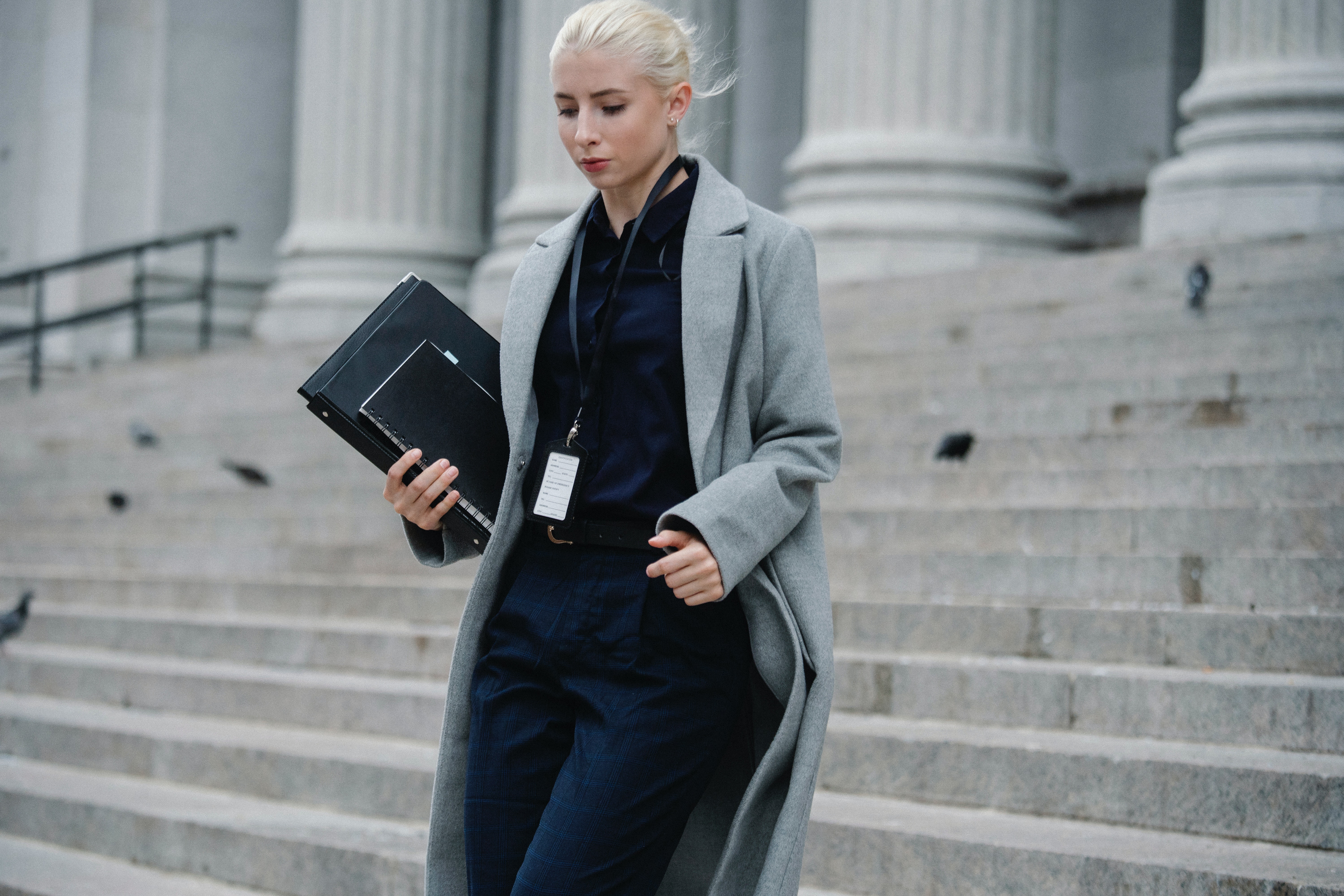 A close up photo of a lady in a lt. gray coat holing a note book and some books, all in black, walking down the steps or what looks like a large courts building.