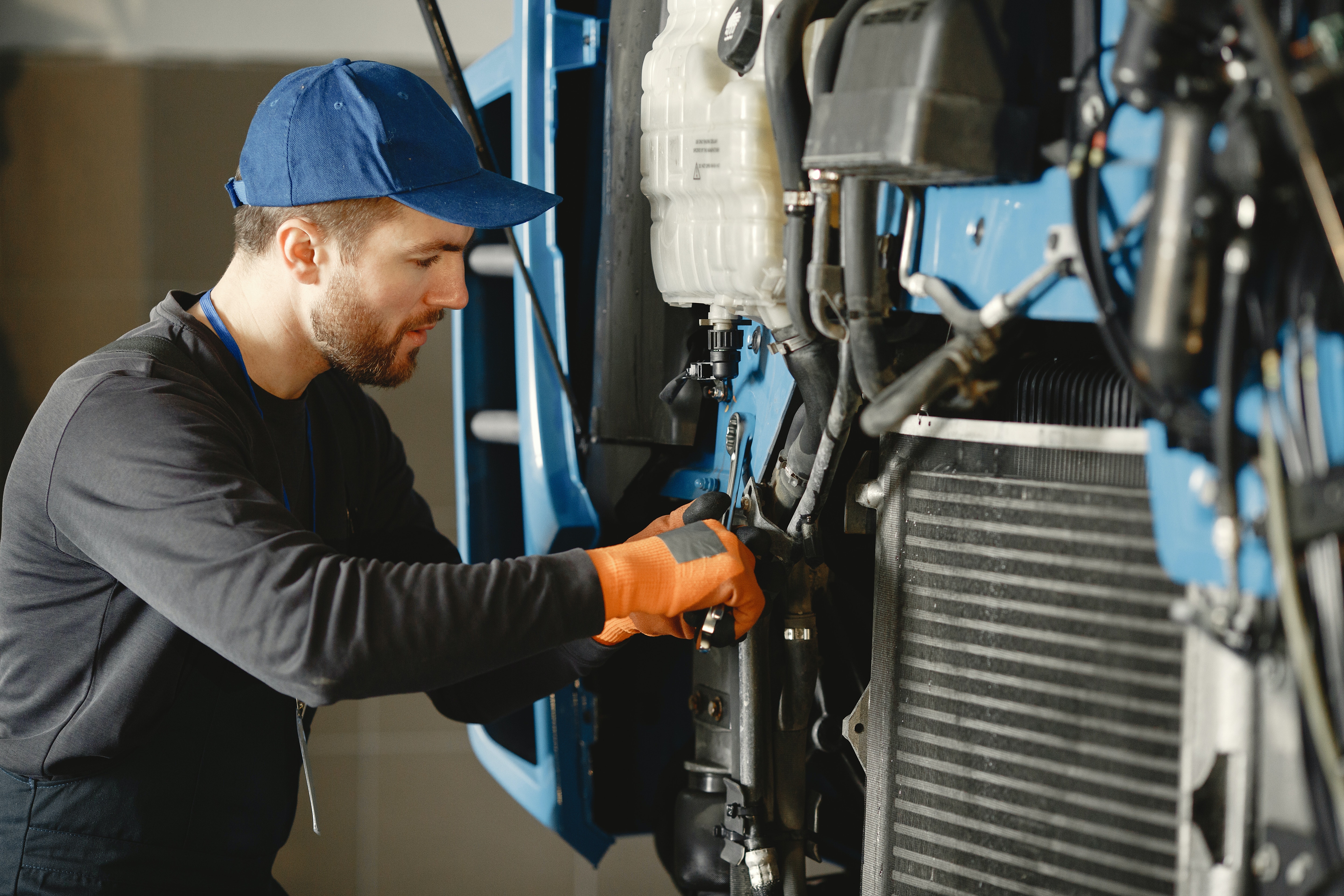 A photo of a man in a gray shirt with a blue cap working on the front of a diesel truck with its hood open with a wrench as seen from the side of the truck's engine.