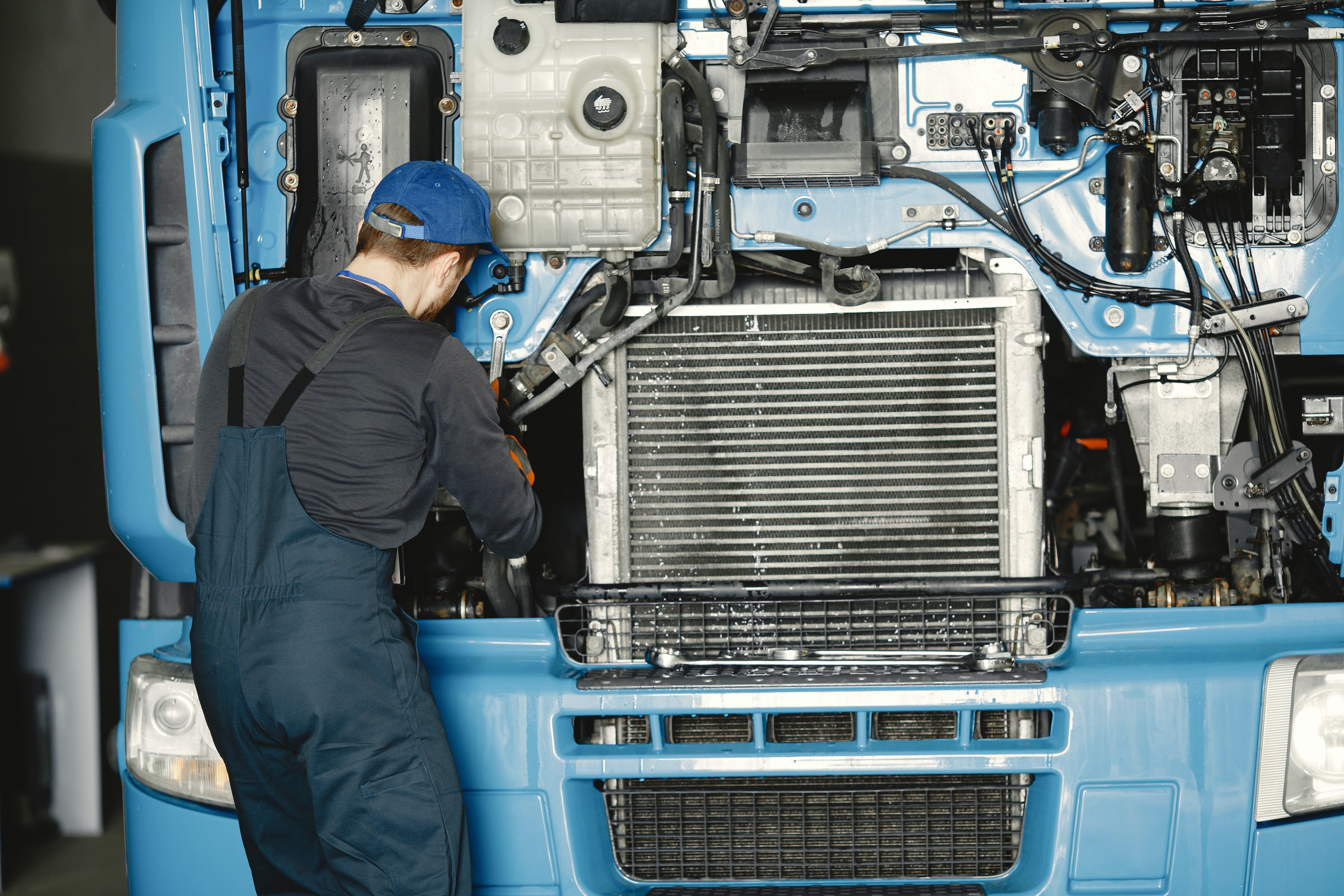 A photo of the same man, from the previous photo, in a gray shirt with a blue cap working on the front of a diesel truck with its hood open turning a bolt with his wrench as seen from the front of the truck's engine.