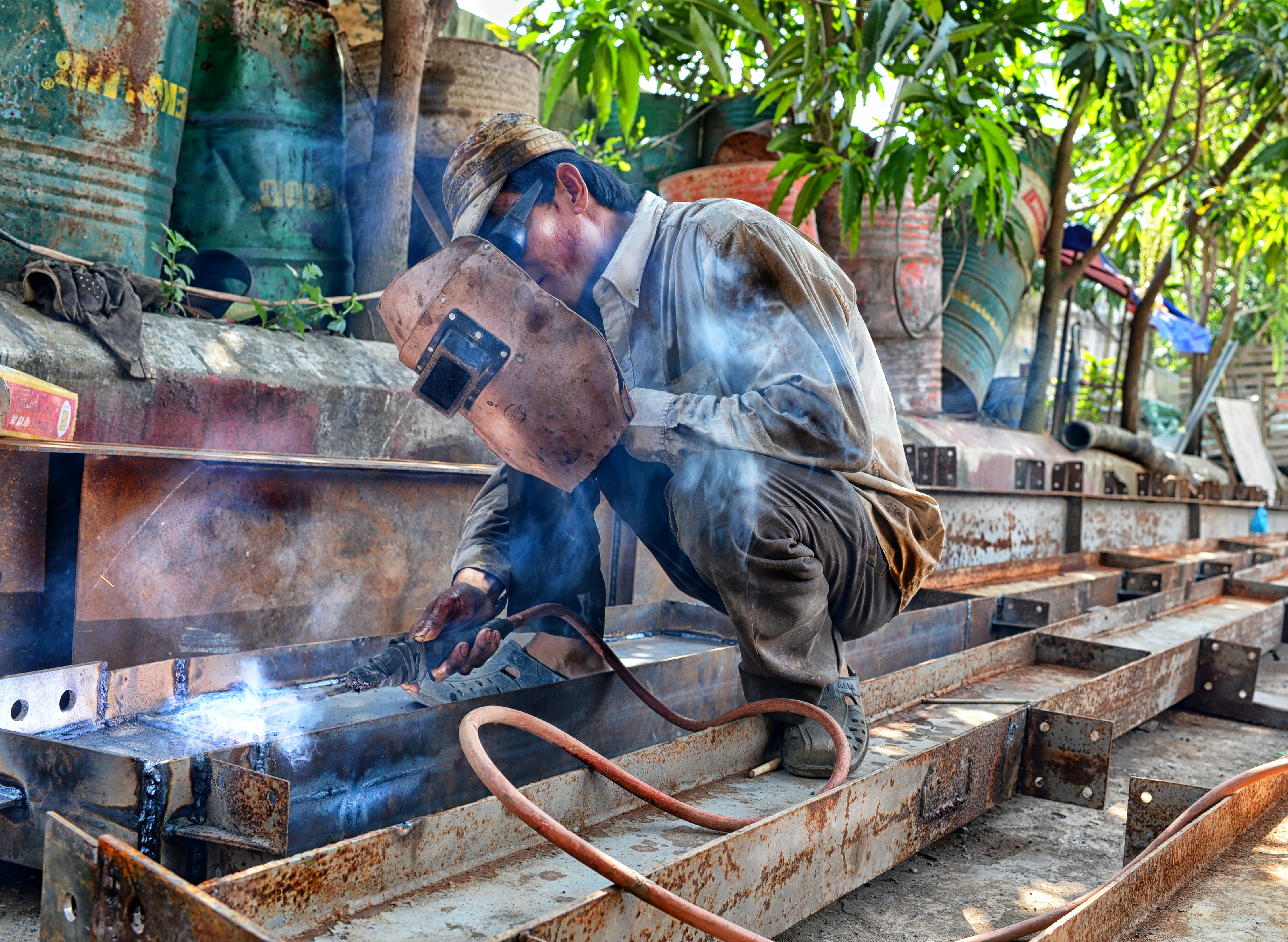 A photo of a man welding outdoors in the shade on some metal beams.