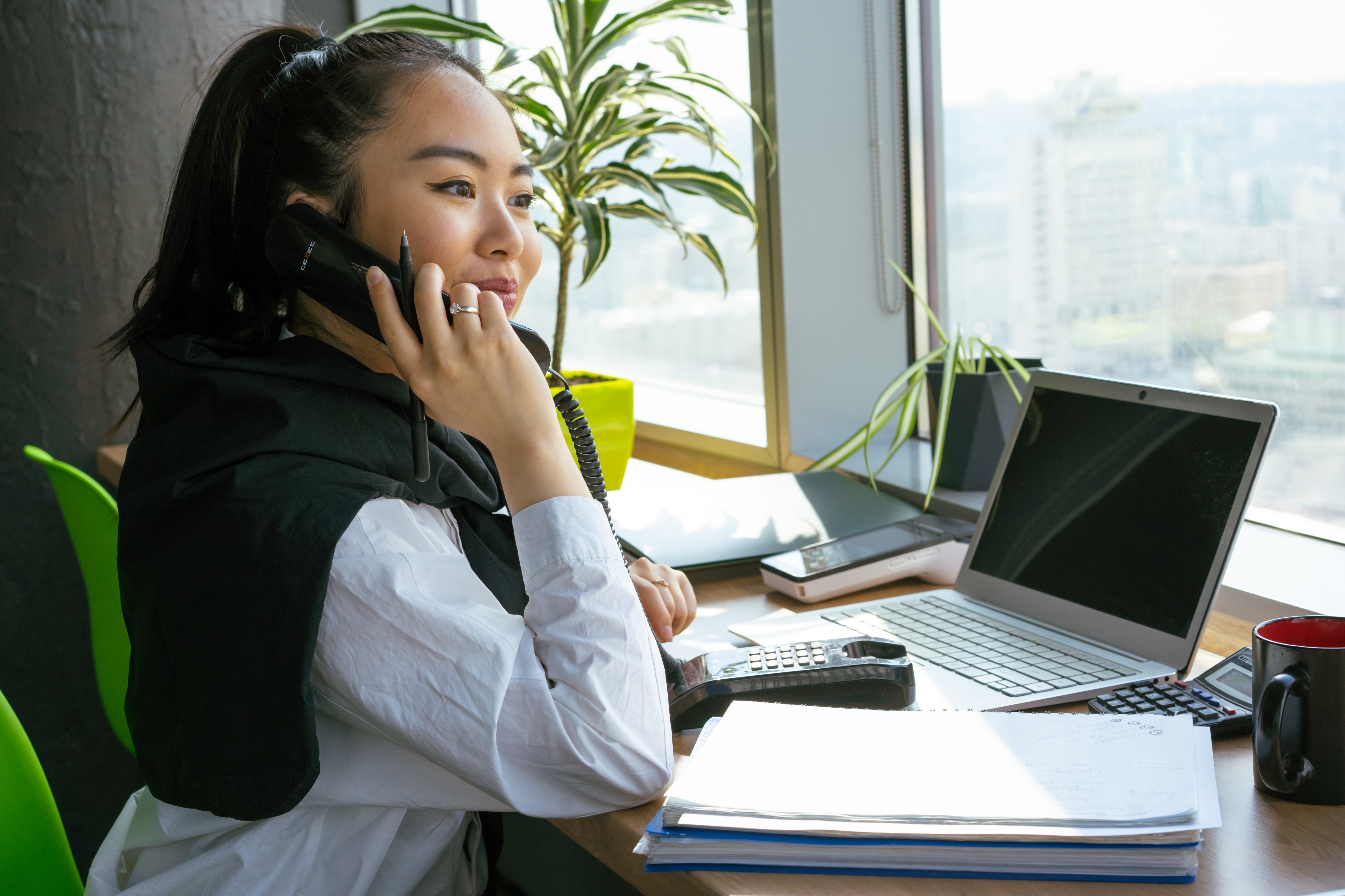 A woman at a work desk, holding a pen and speaking on a phone.