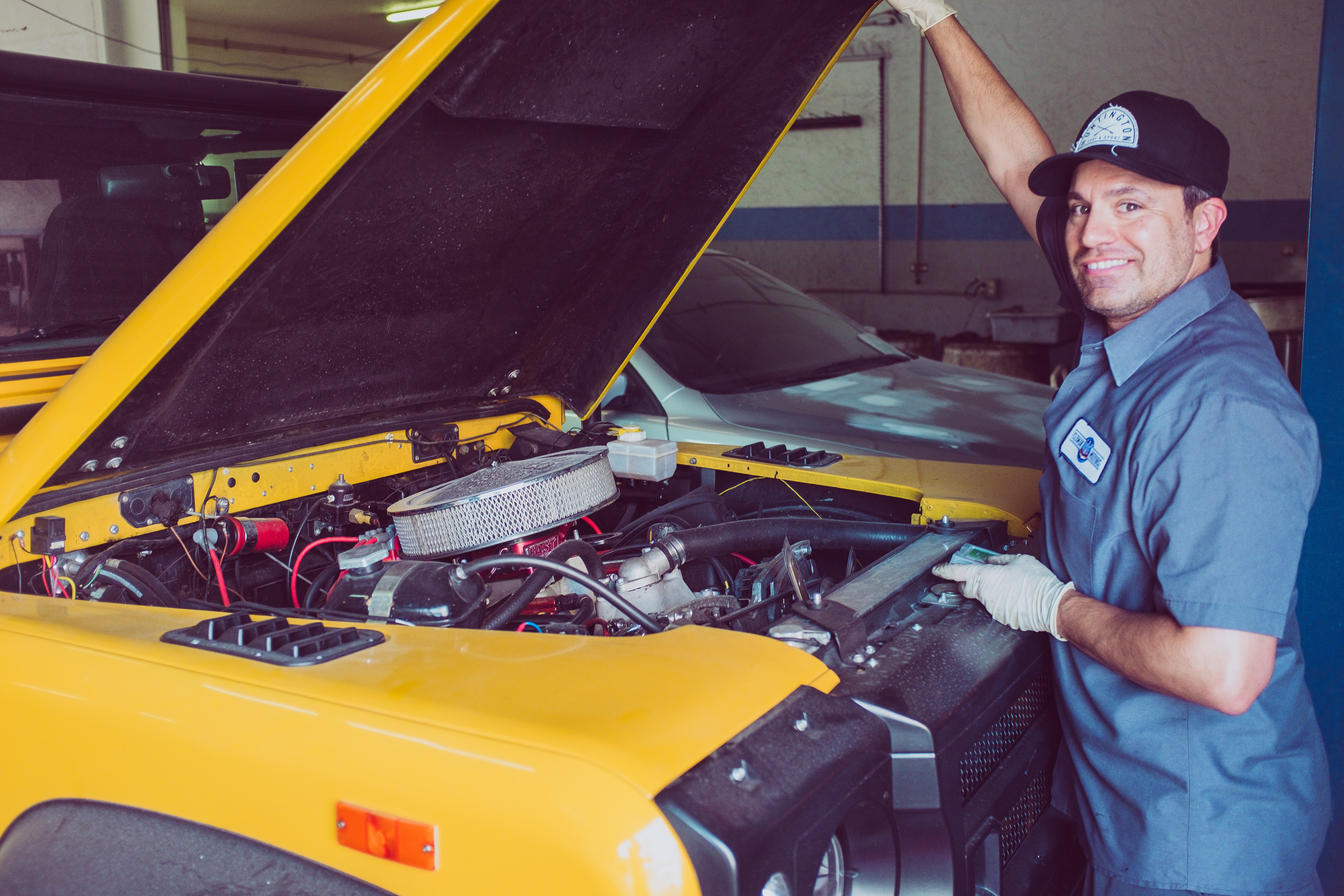 A medium closeup photo of a mechanic, in a garage, standing and holding up the hood of a yellow, car, SUV or truck while smiling at the camera. The top of the engine is showing and most notably its air filter can be seen.