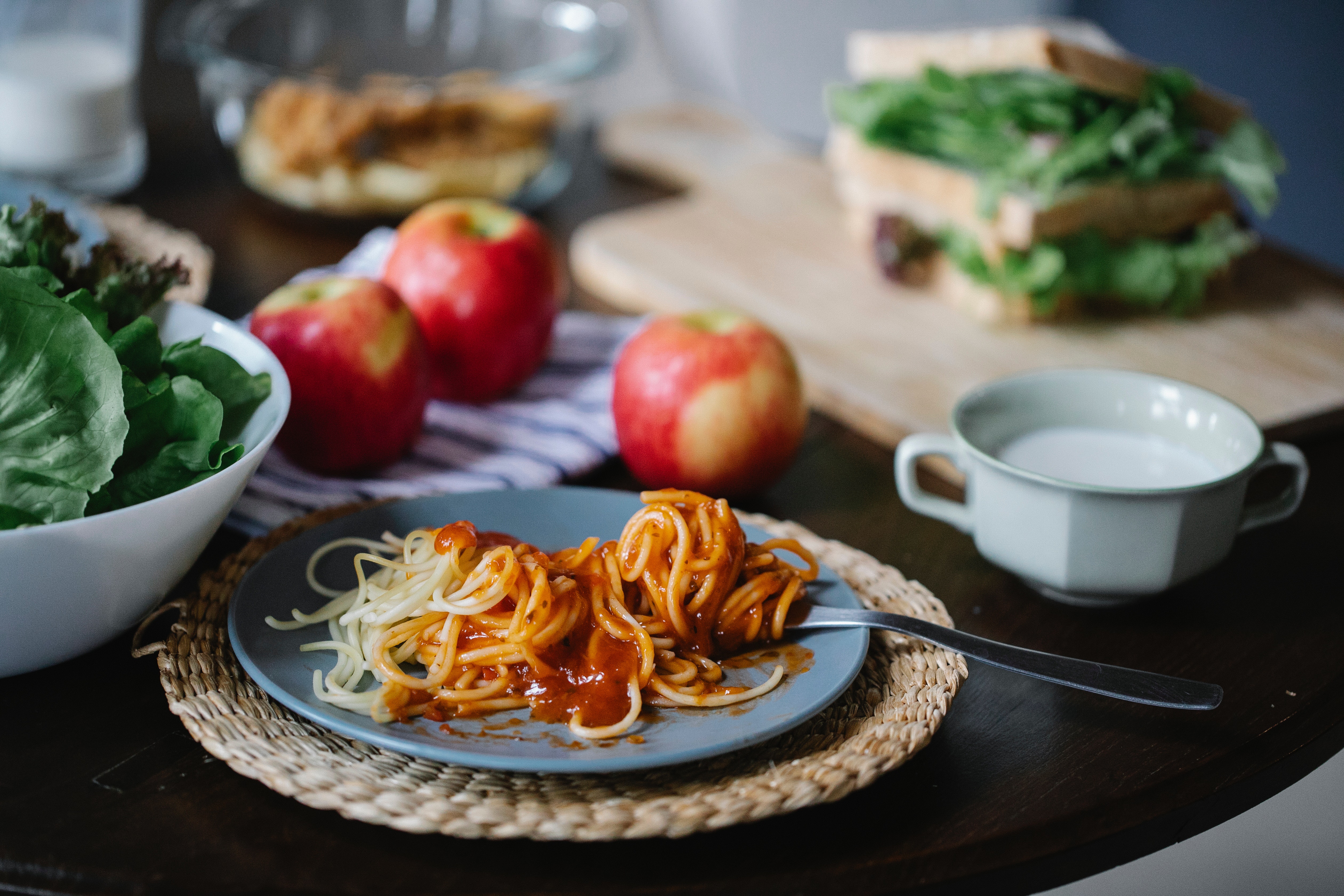 A photo of a plate of delicious spaghetti with a salad, an apple and a cup of soup or cream.