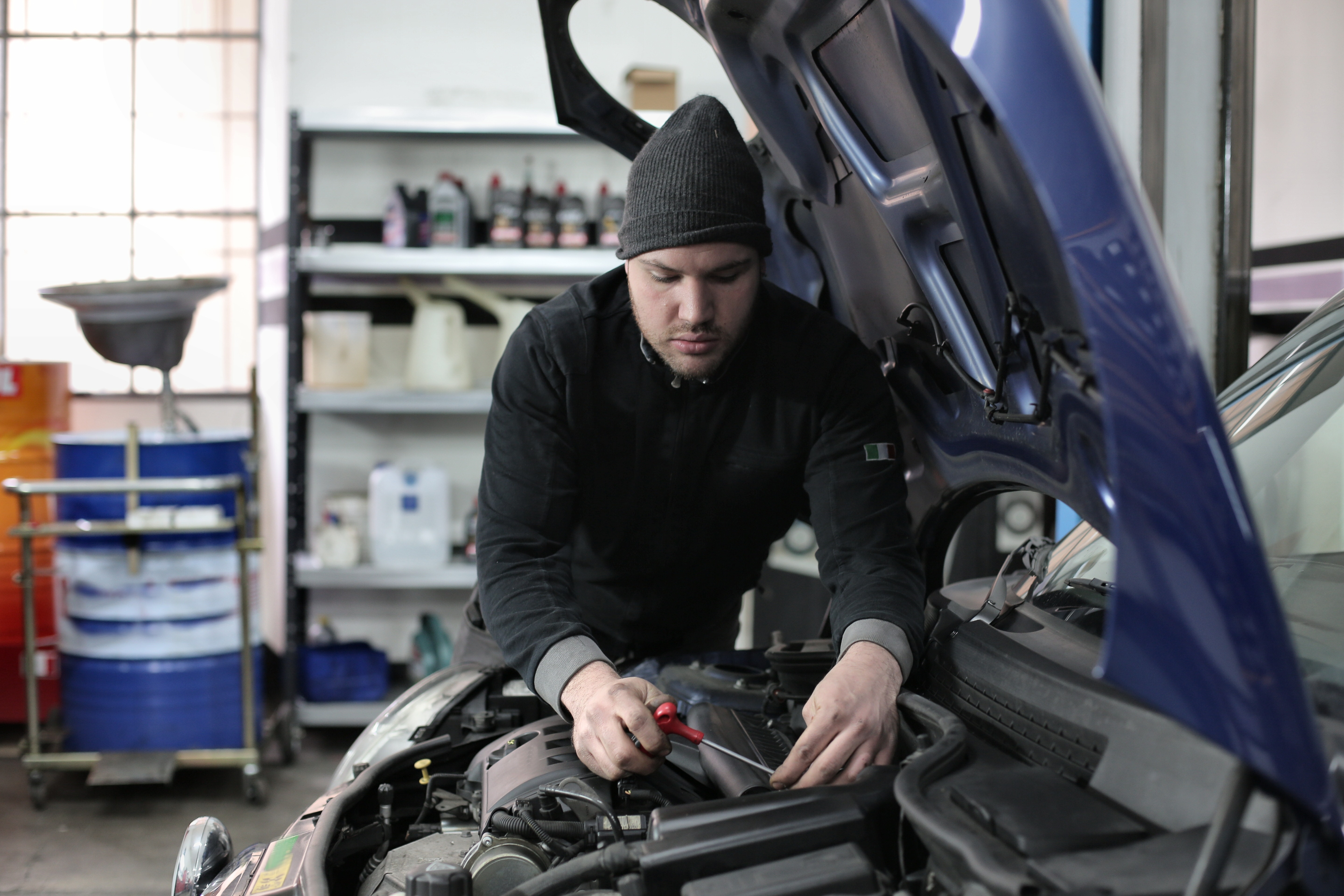 A medium closeup photo of a mechanic, in a garage, working under the hood of a medium, blue small car, taken from the side.