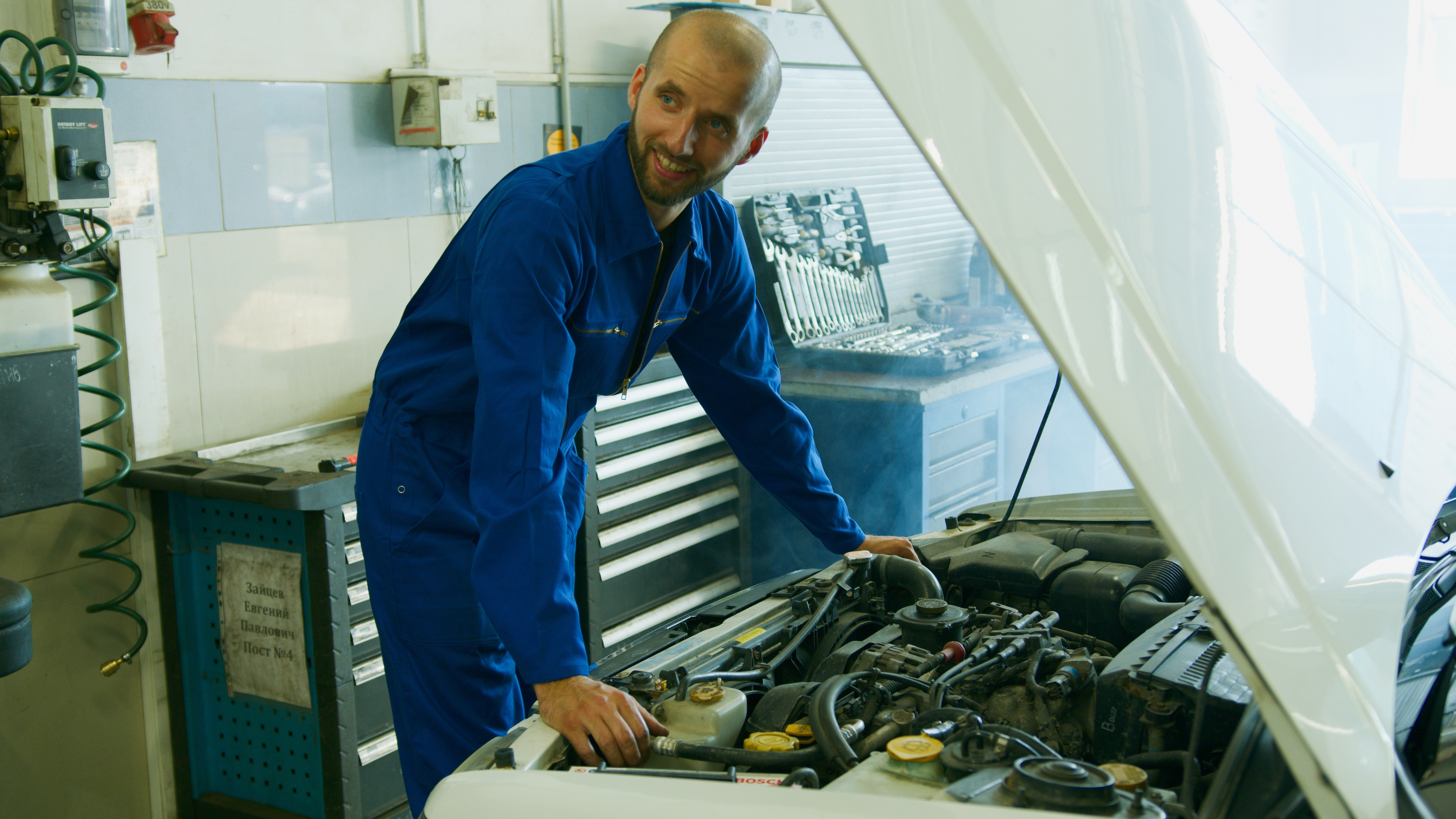 A man with blue coveralls looking to the right while working on a car's engine under the hood.