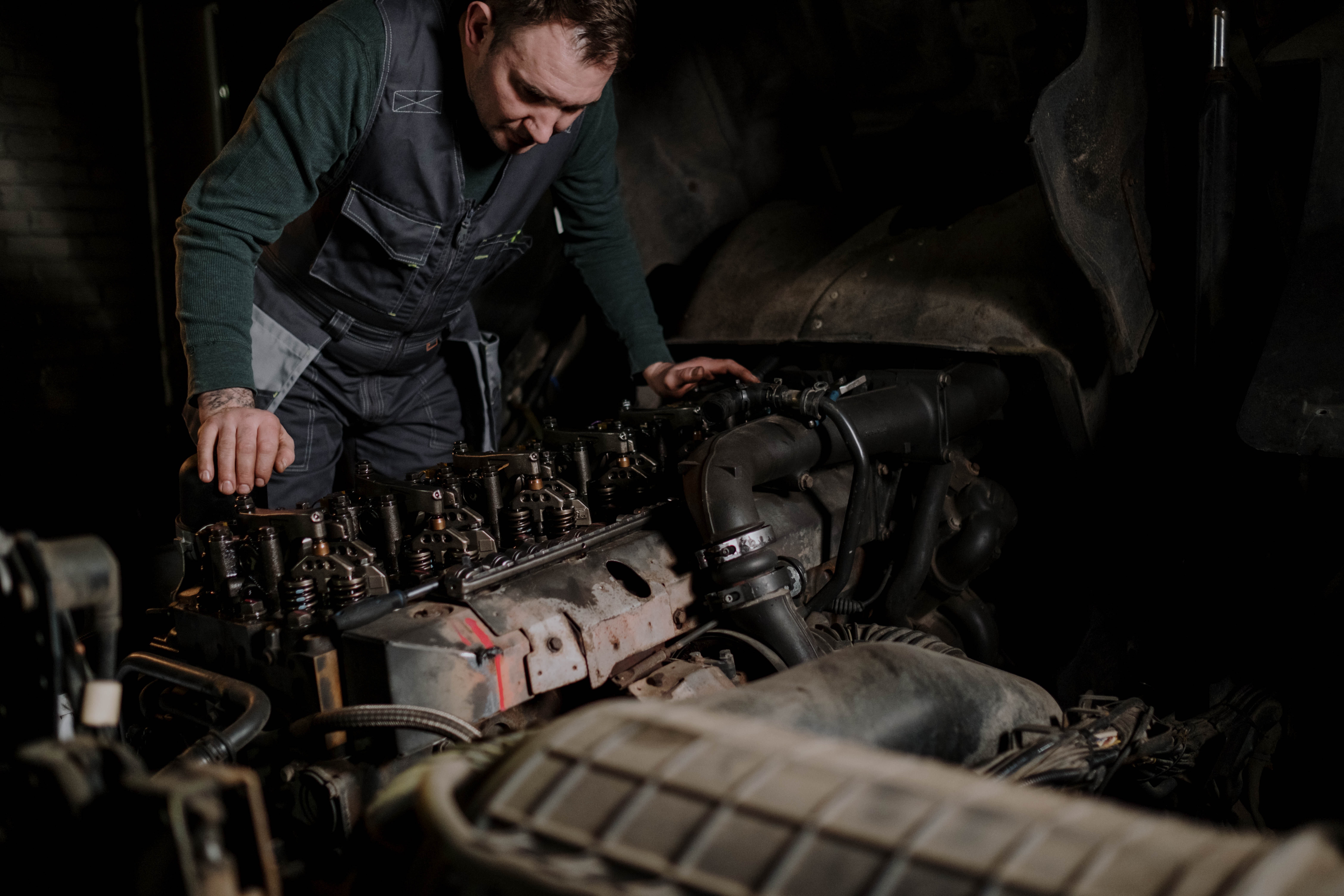 A photo of the same diesel mechanic working on the same engine, from the previous photo taken facing the engine, working alone with the hood open.