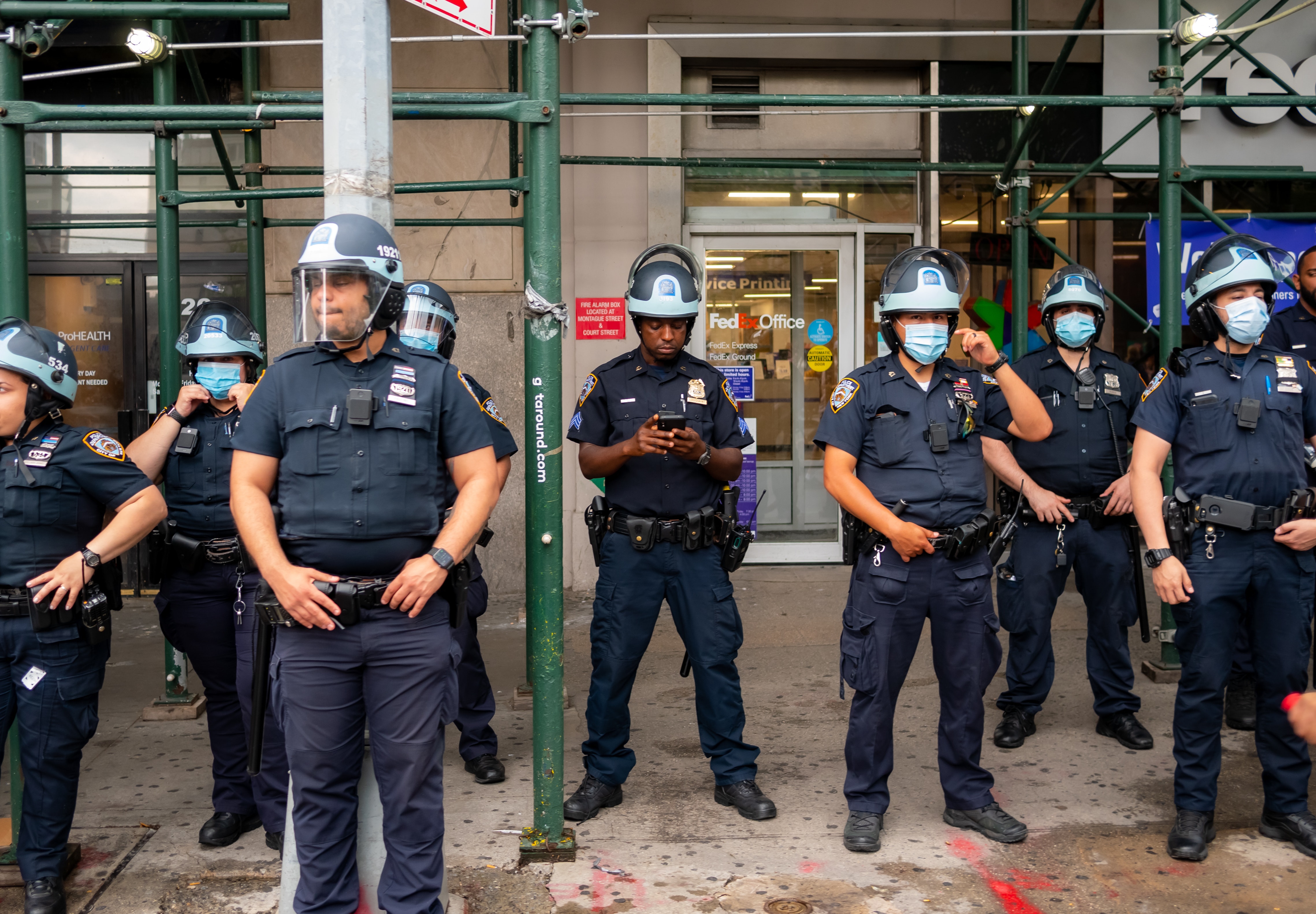 A photo of a loose line up of 8 policemen wearing surgical masks in front of some businesses. One of the policeman is looking at a hand held device with his mask off.