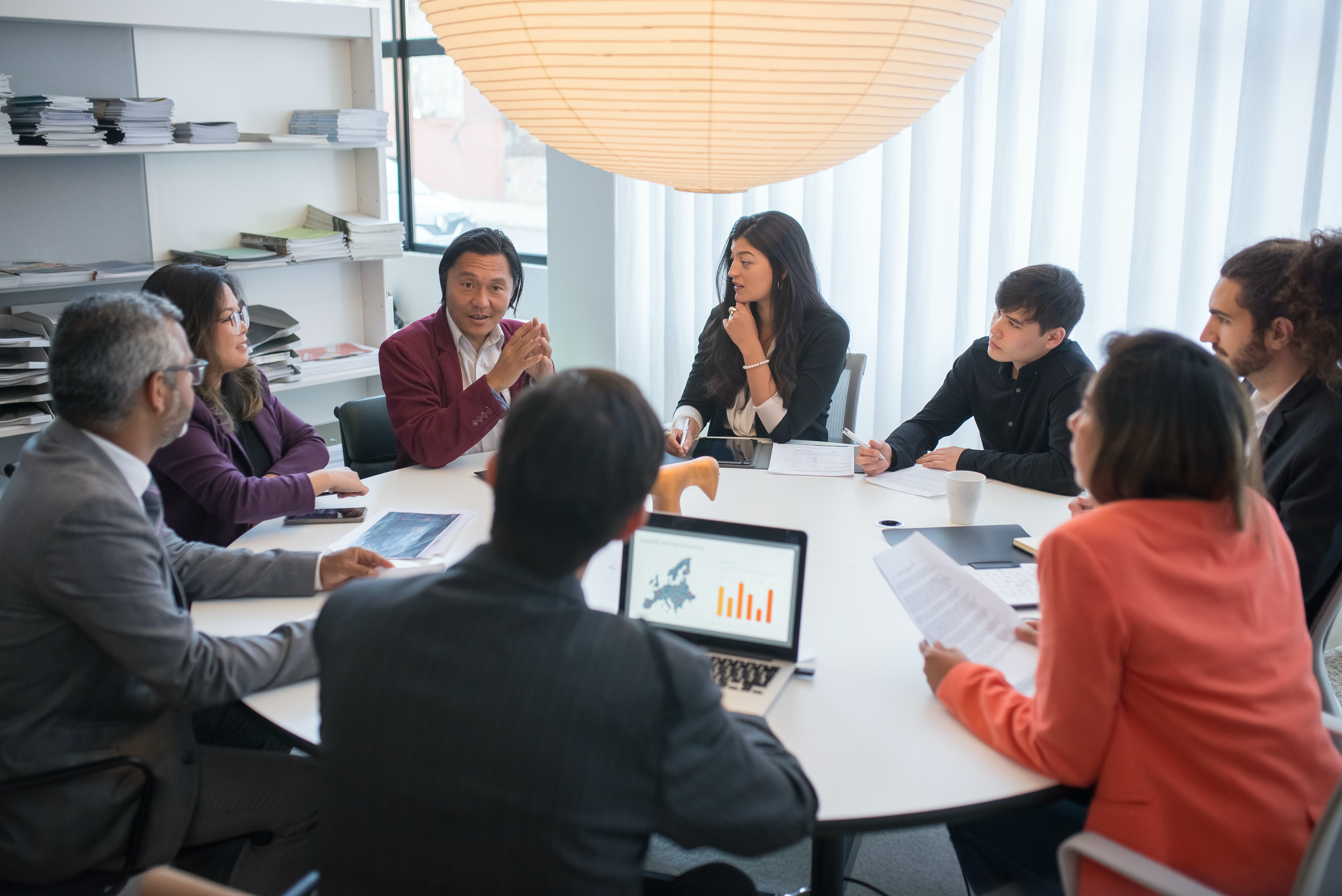 A photo of a group of 8 people in an office setting at a business meeting being lead by their manager. This appears to be a daytime meeting and room setting is predominantly bright white with the sunlight brightening up the white drapes covering a large window.