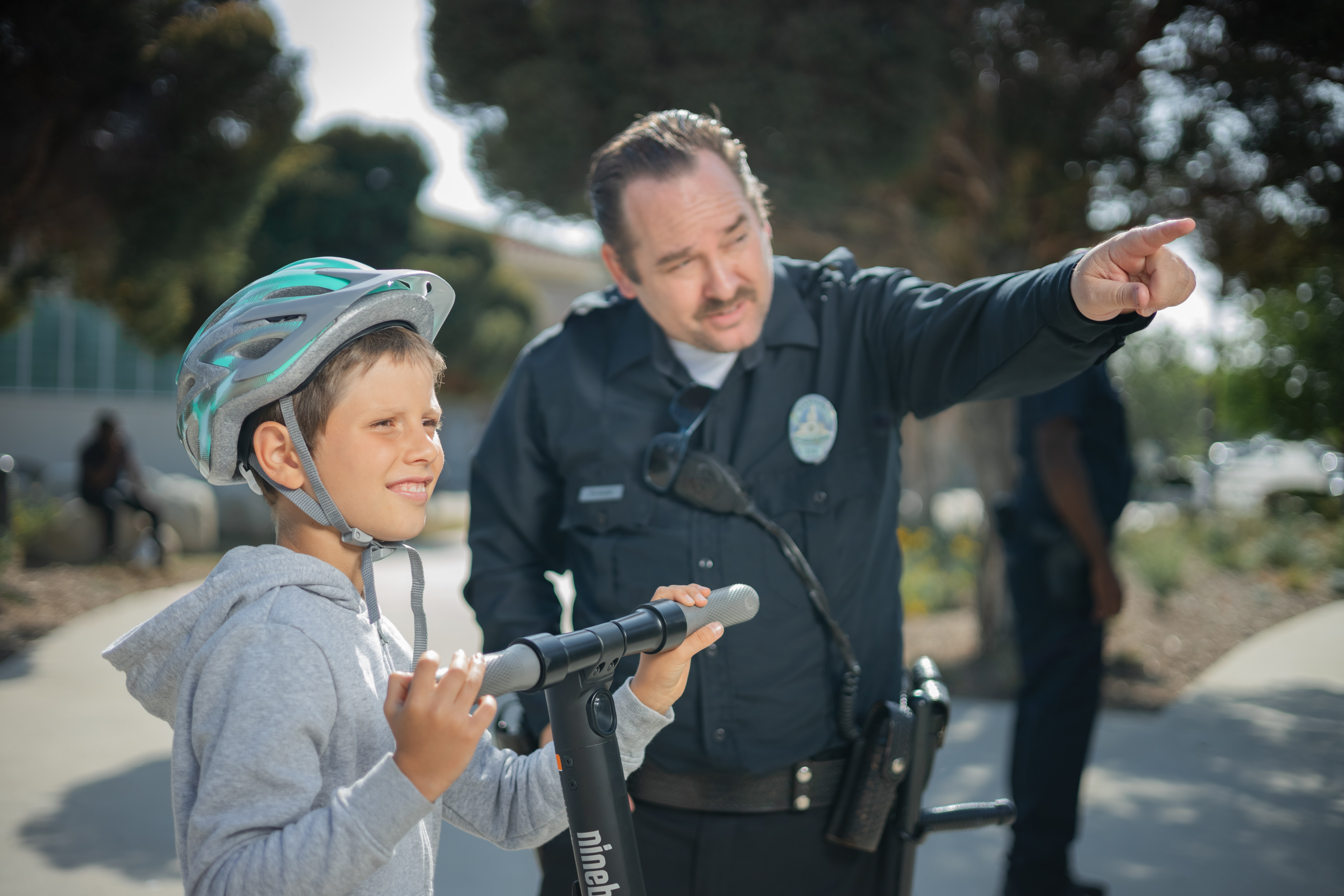 A medium closeup photo of a policeman pointing to show a boy on a scooter something off in the distance.