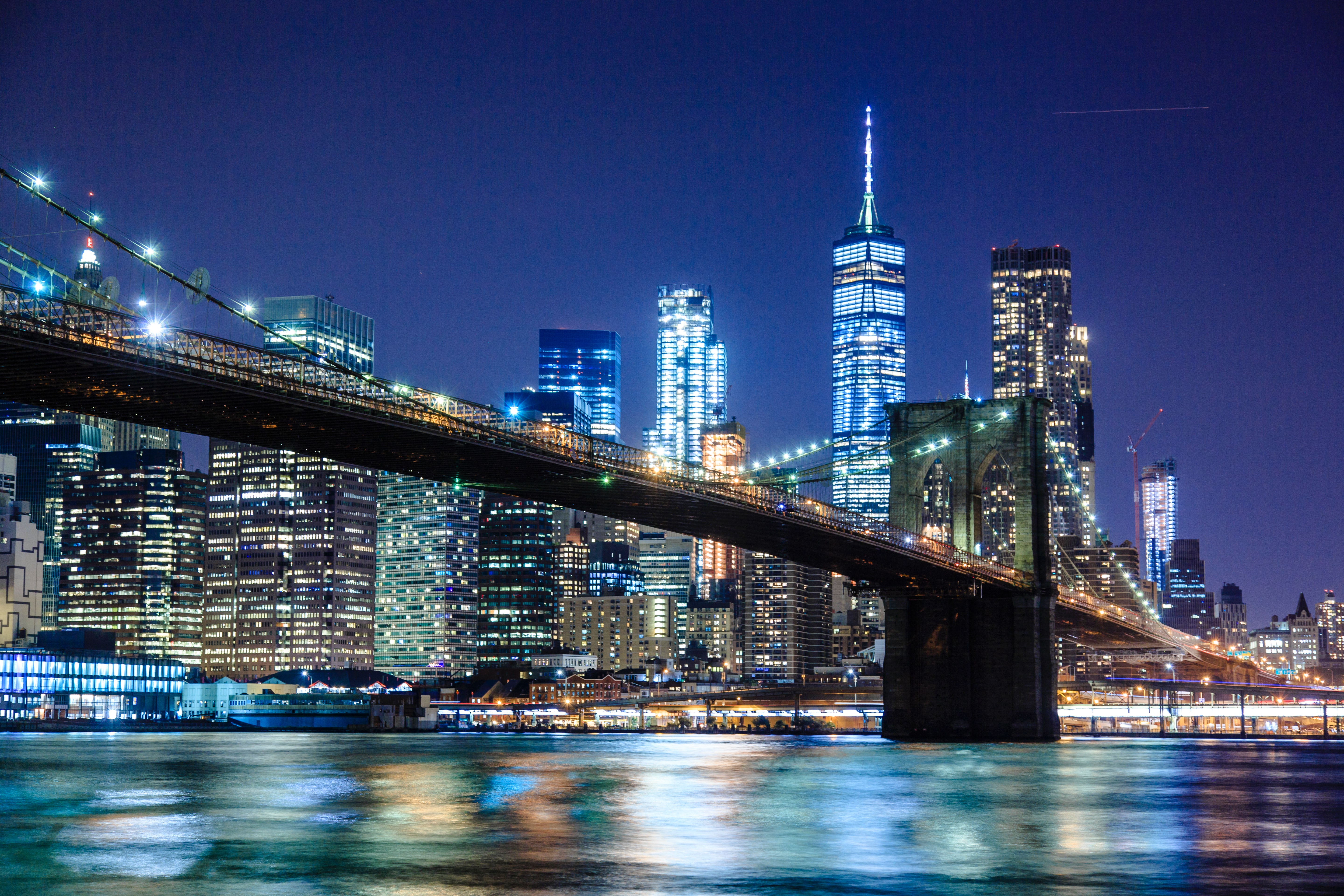 A night photo of the Brooklyn Bridge against the brilliant, deep blue New York city skyline with all the cities lights glowing behind it.