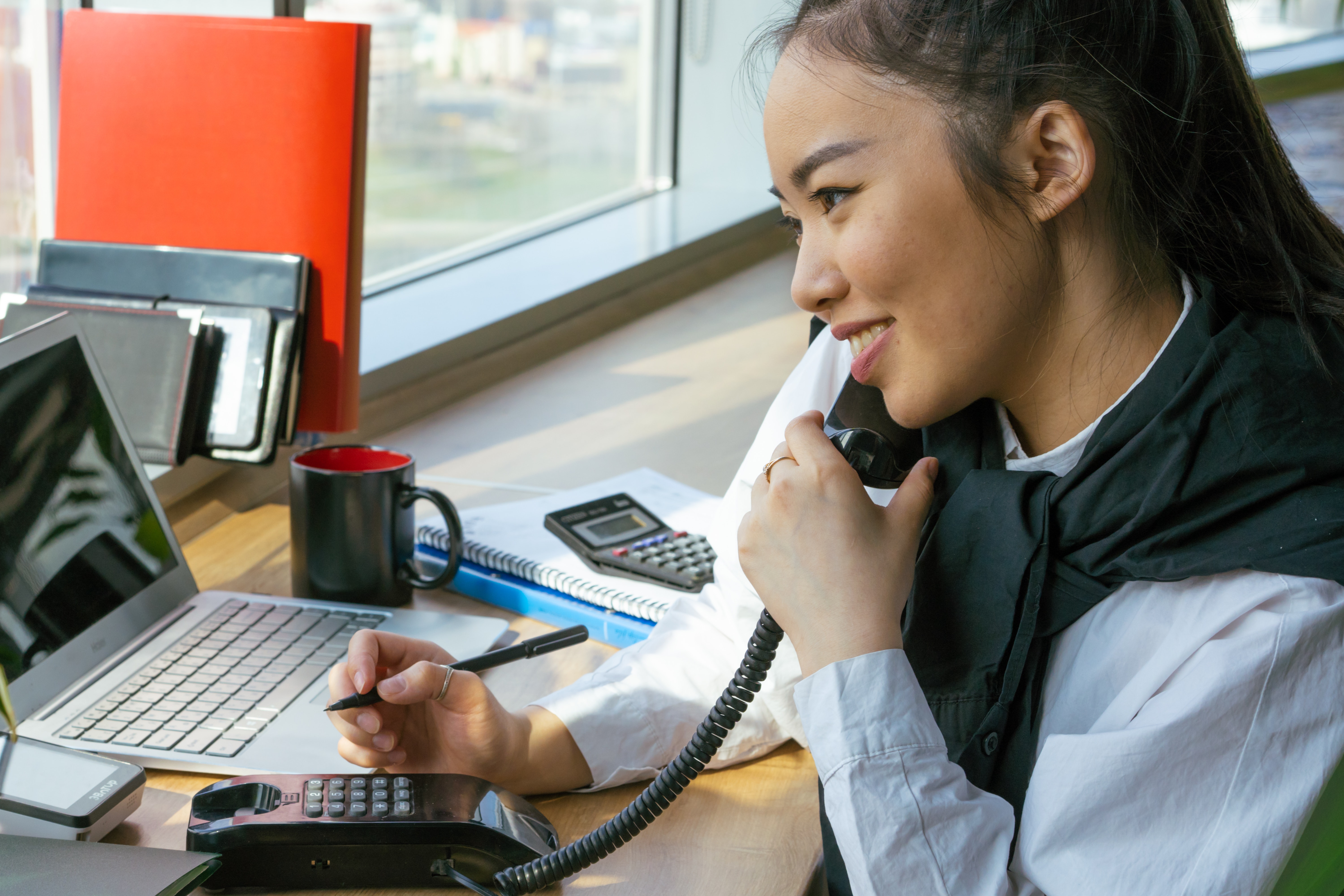 A photo of an accountant at her desk on the phone smiling in front of a window with her laptop computer, notpads and calculators in front of her.