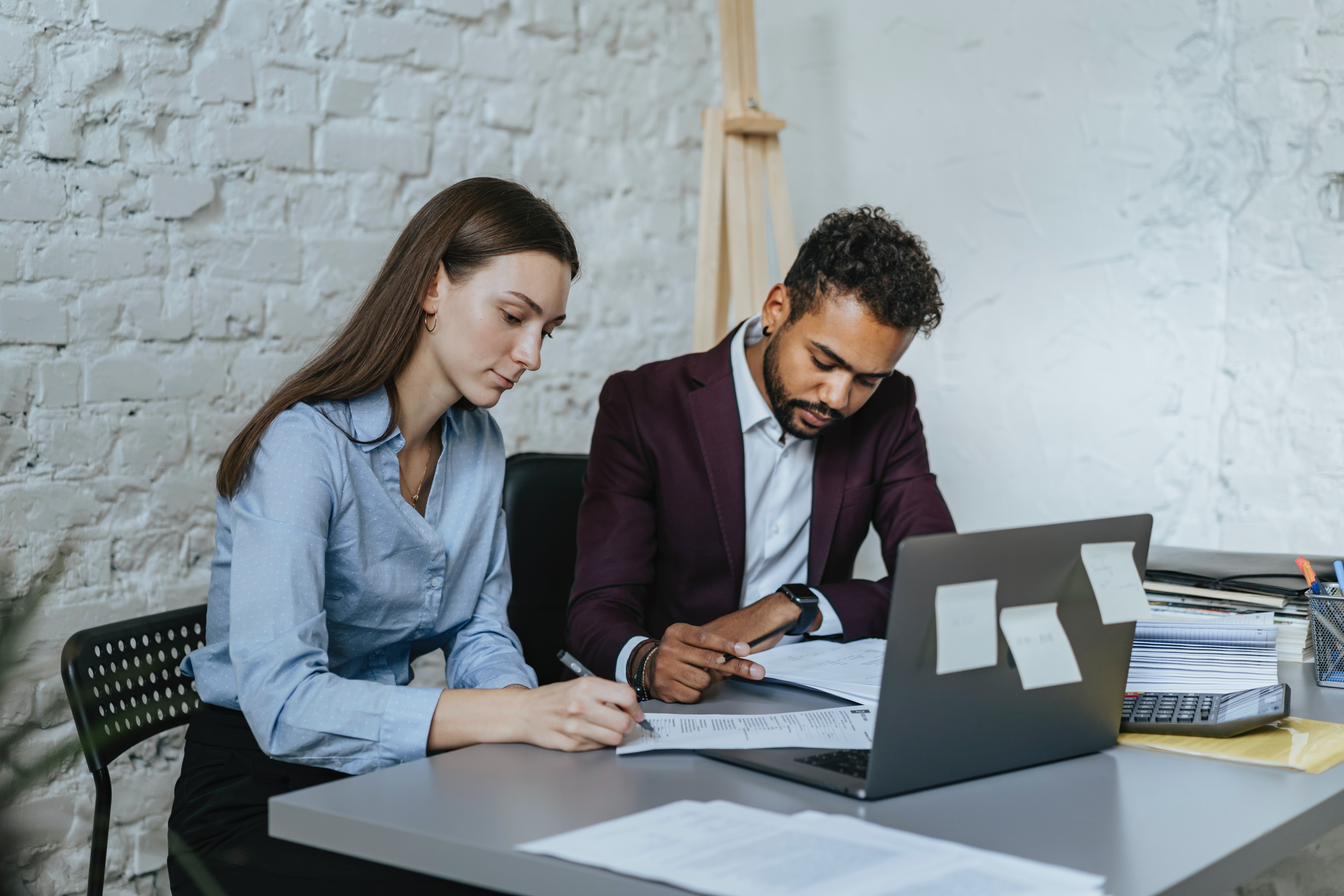 A photo of a man and woman, who may be paralegals, sitting at desk in front of a laptop computer going over paperwork. They're in a bright office with brick walls painted white.