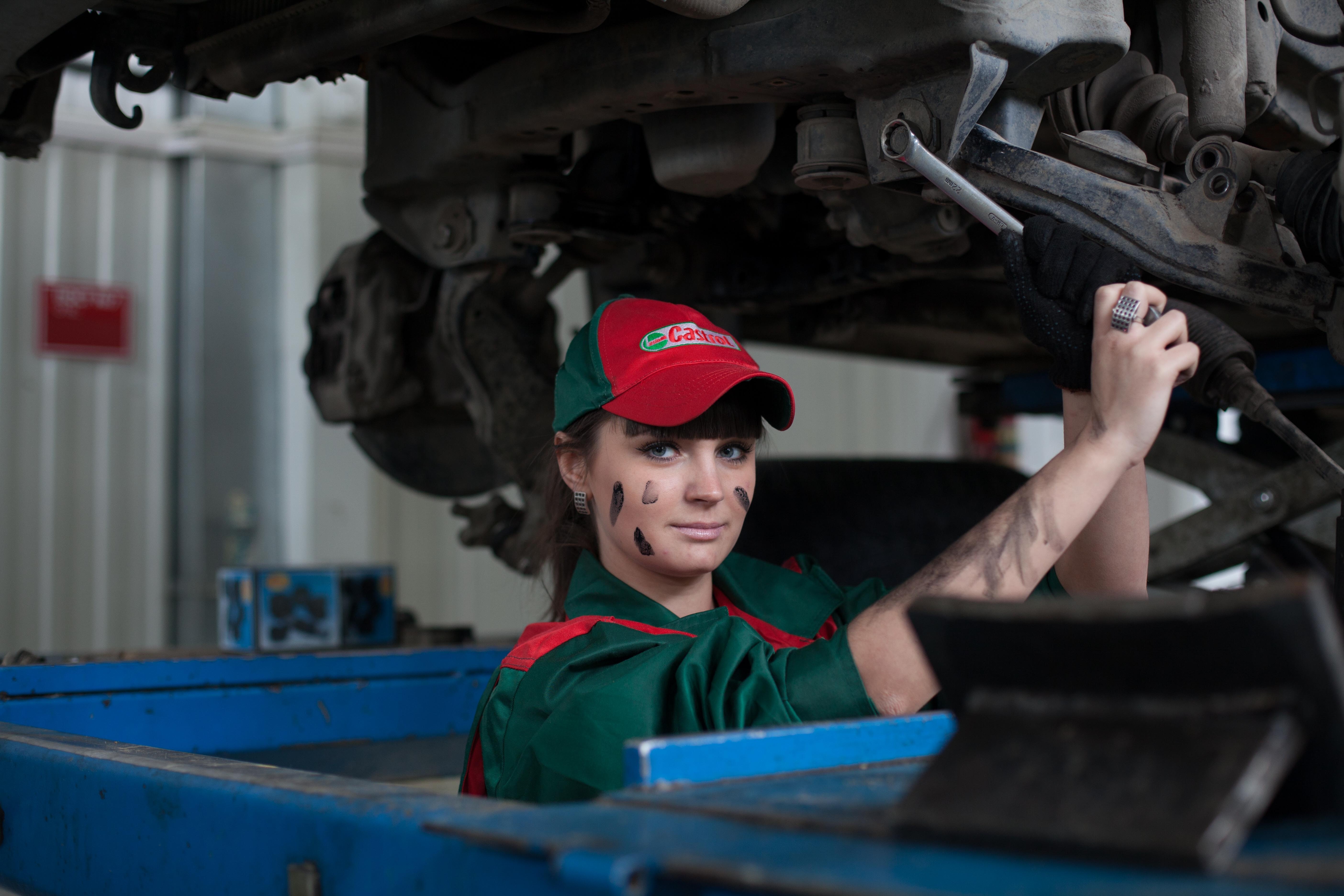 A photo of a young lady working underneath a diesel truck dressed in green and red with a company baseball cap on looking at the camera with a light smile on her face.