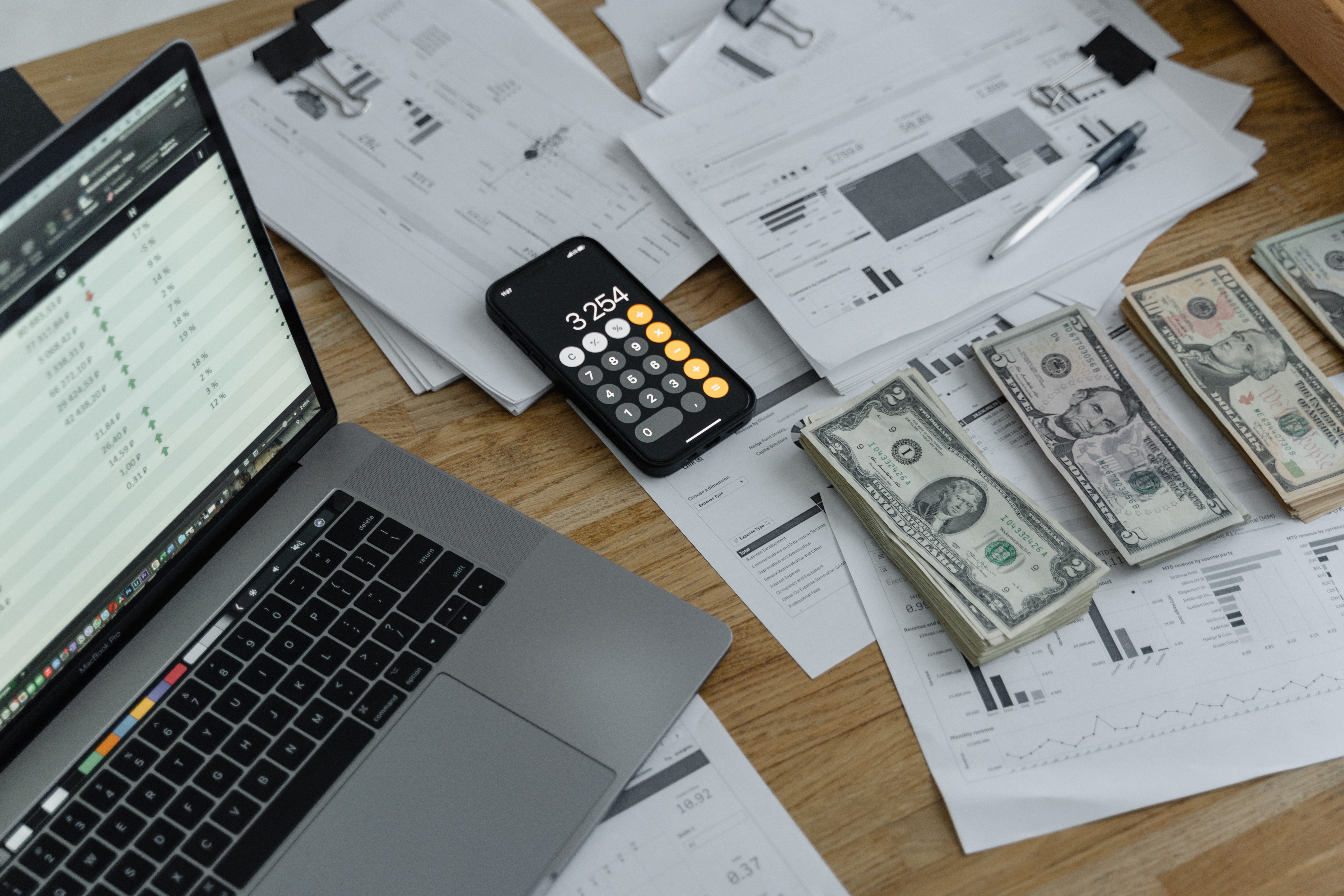 A photo a laptop computer, a calculator, papers and stacks of money on top of a table.