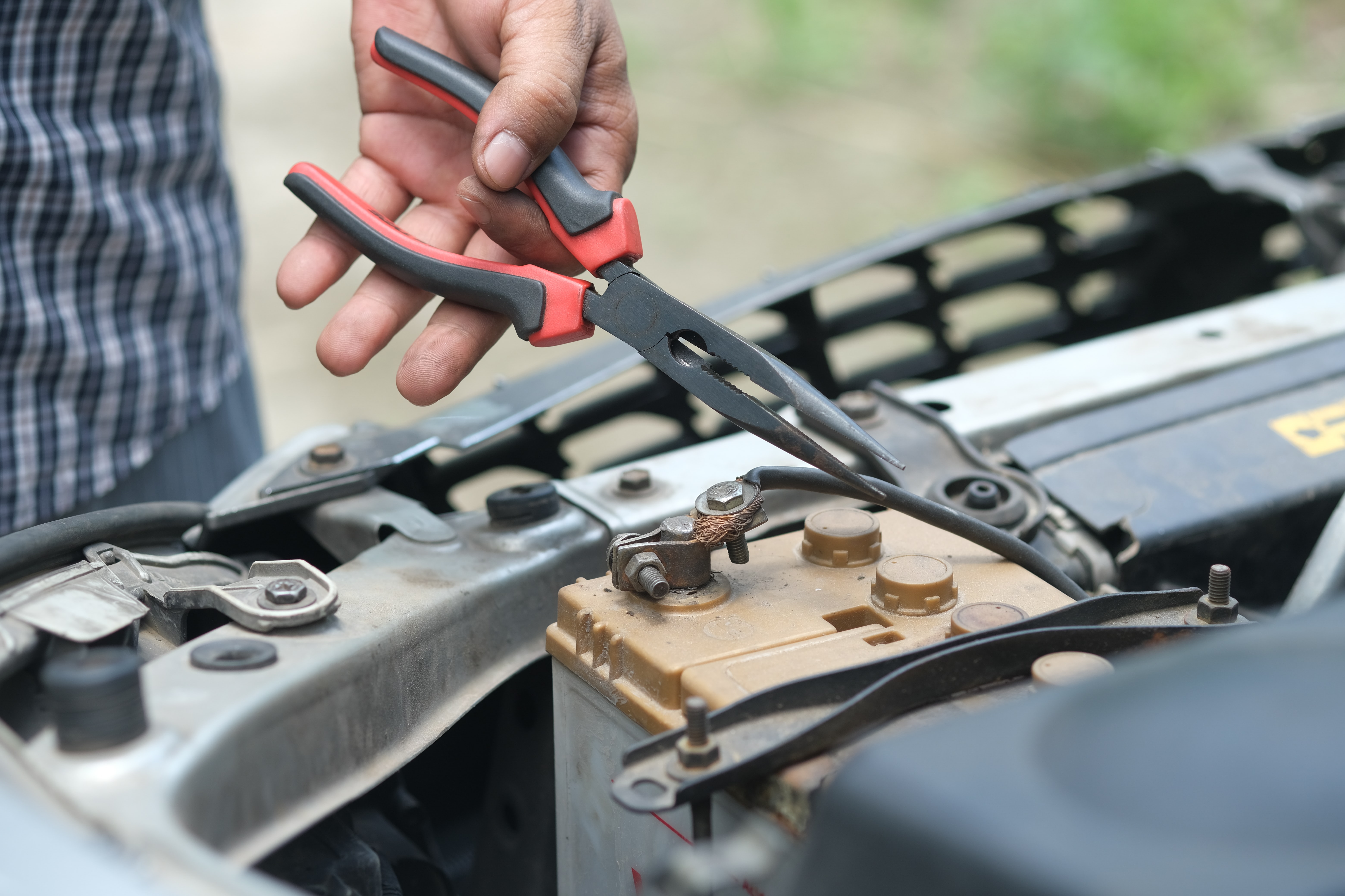 A close up of a person's hand with a pair of needle nose plyers about ready to pull a black wire attached to a car battery.