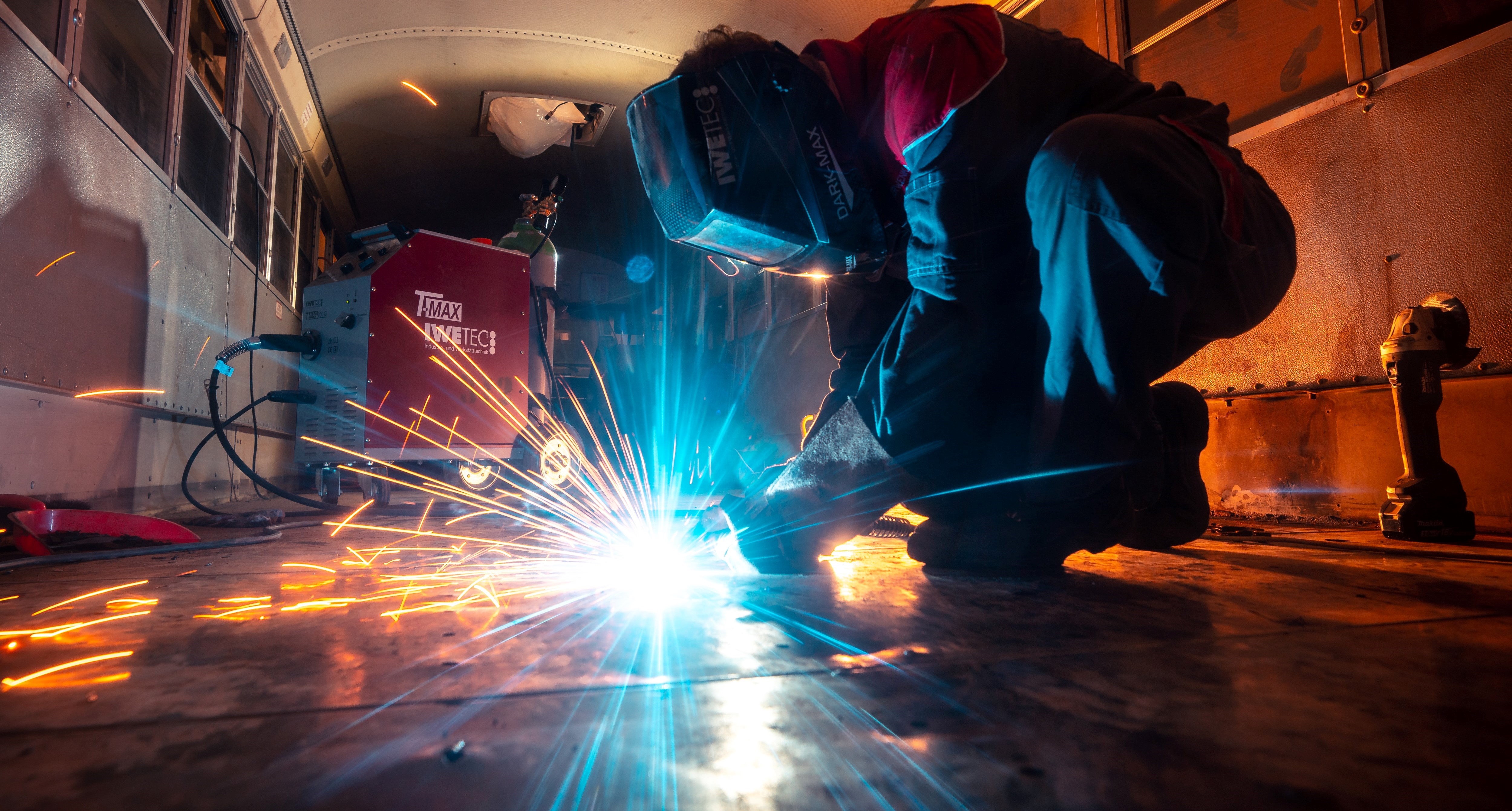 A person in blue coveralls and a mask, kneeling and welding on what looks like the floor of school bus.