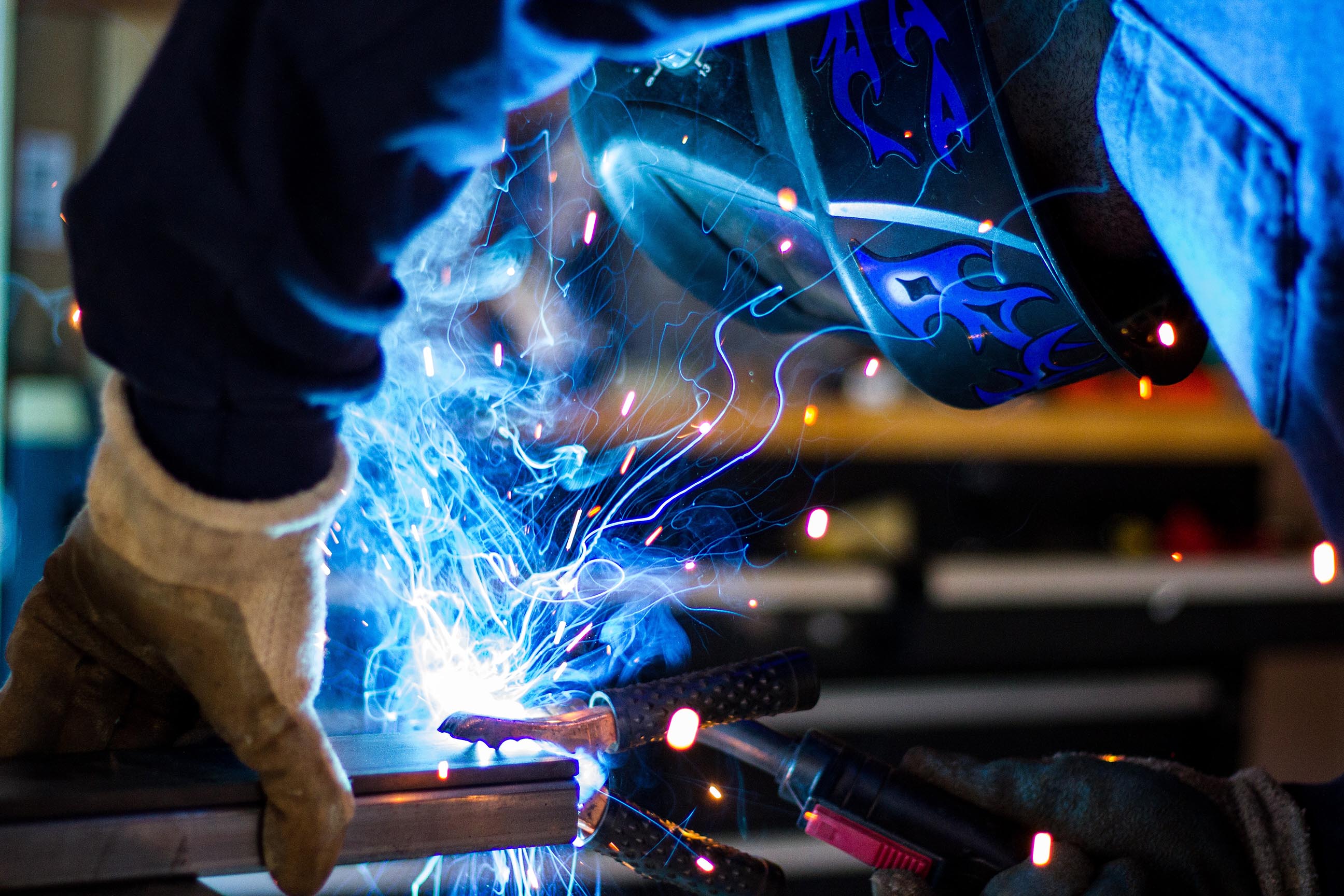 A close up photo of a person dressed in blue coveralls welding on what looks like to be the end of some rectangular tubing about 3 – 4 inhes wide and quite long.